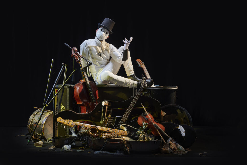 Personaje teatral con máscara blanca y sombrero entre instrumentos de música