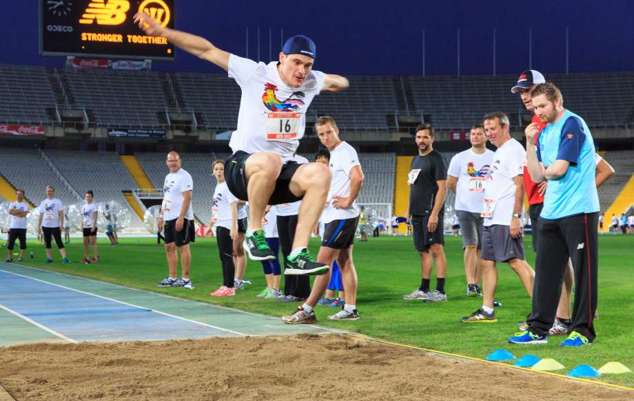 Participante realizando salto de longitud durante actividad de team building deportivo en estadio olímpico por la noche con compañeros animando bajo el lema Stronger Together