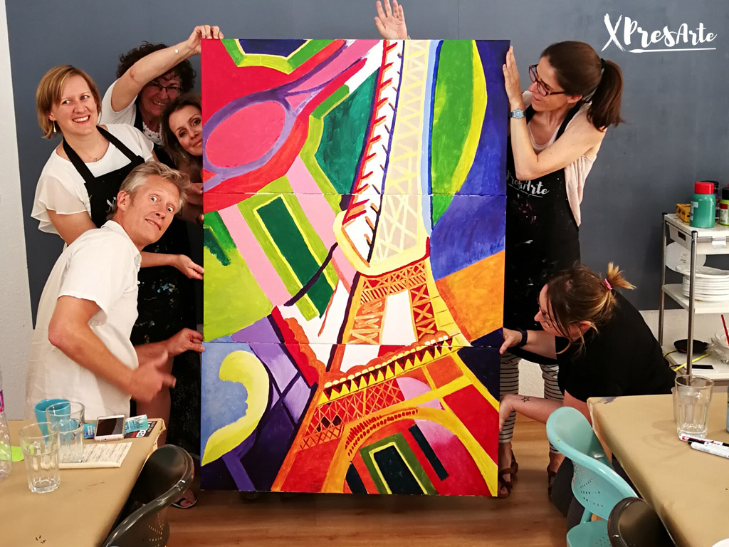 Equipo de compañeros mostrando orgullosos un mural colaborativo de la Torre Eiffel pintado durante actividad de team building artístico con XPresArte