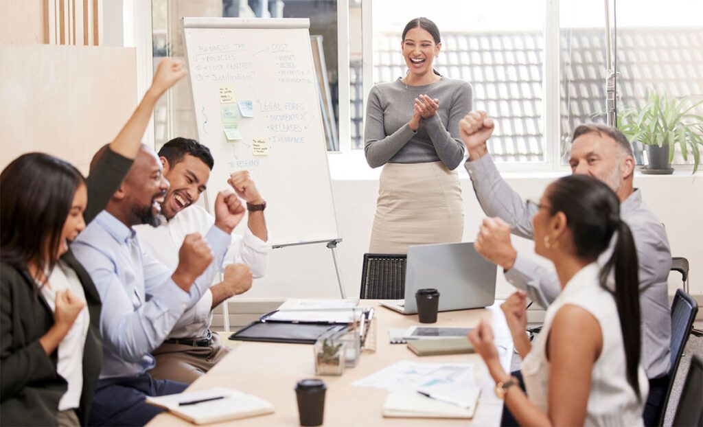 Equipo celebrando un logro durante una dinámica de motivación, con una facilitadora aplaudiendo y pizarra con notas al fondo