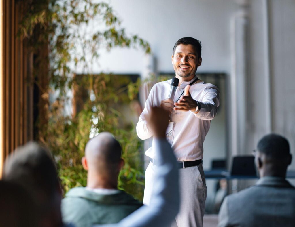 Ponente masculino con micrófono señalando a asistente con mano alzada durante sesión de preguntas en conferencia corporativa en espacio moderno con vegetación natural de fondo