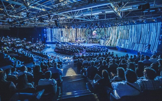 Gran auditorio con aforo completo de profesionales durante fiesta de empresa de gran formato con escenario inmersivo de pantallas LED azules y espectáculo en directo
