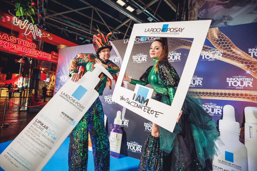 Dos intérpretes con vestuario brillante posan con un marco que dice ‘I AM #SKINHEROE’ frente al photocall ‘LA ROCHE‑POSAY INNOVATION TOUR 2024’, con botellas gigantes de producto; neón ‘WAH’ al fondo