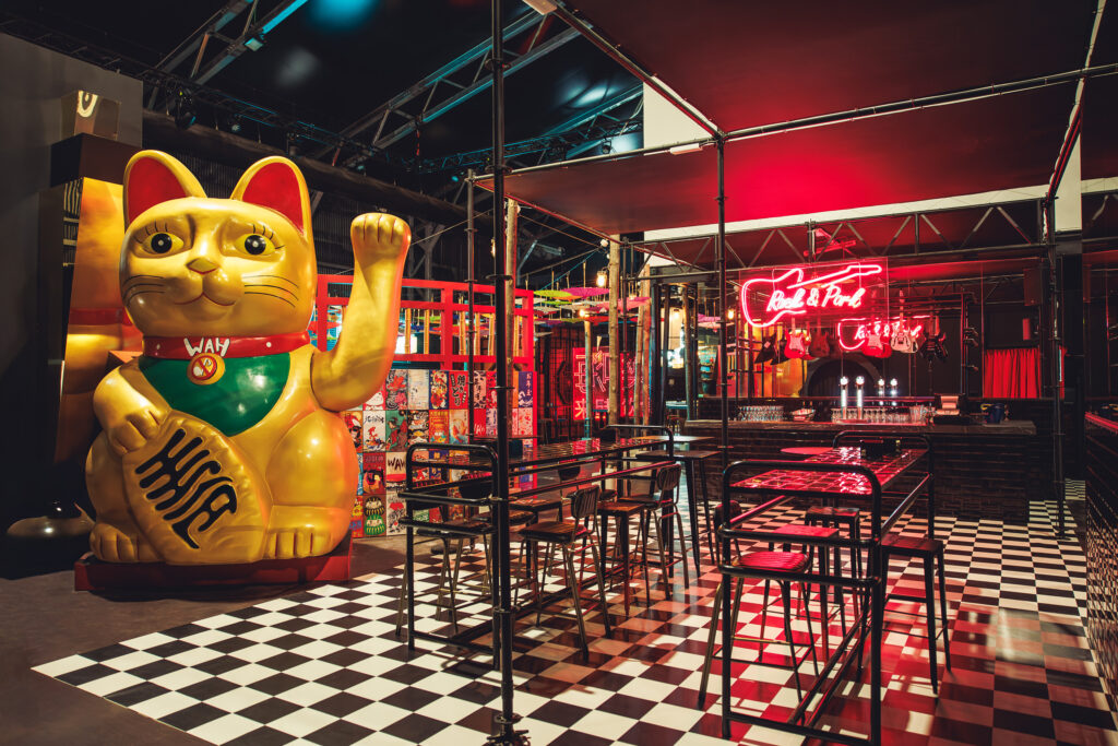 Bar de WAH con maneki‑neko dorado gigante, mesas altas y neón rojo en forma de guitarra sobre la barra, suelo ajedrezado