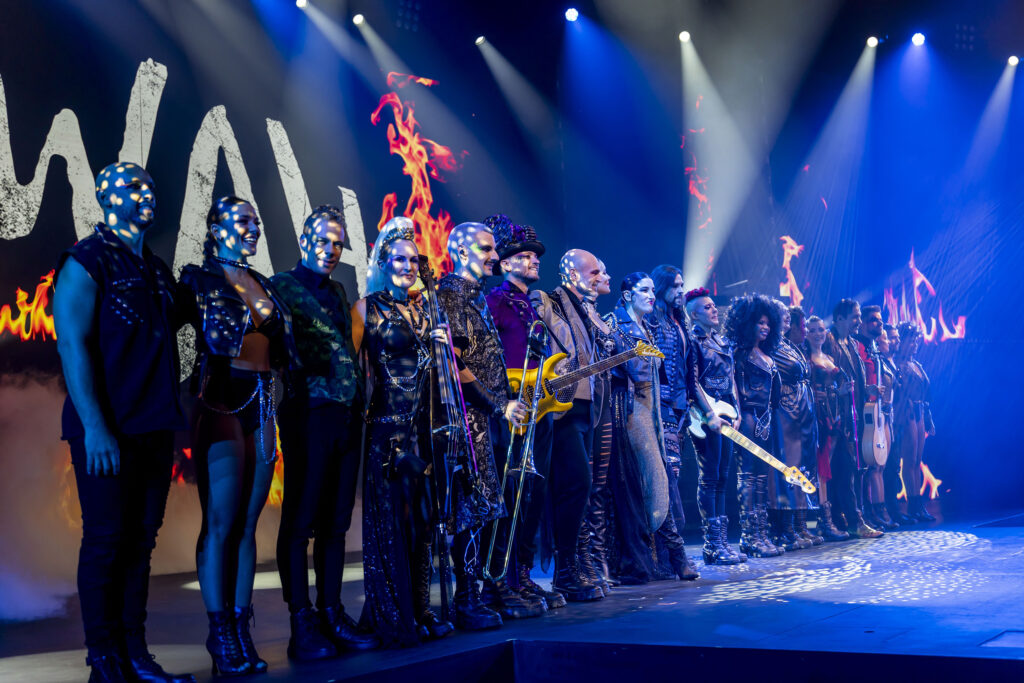 Reparto de WAH Show en línea sobre el escenario con instrumentos musicales, luces azules y el logo WAH detrás.