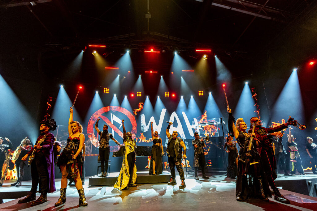 Elenco en el escenario con el logo ‘WAH’ y símbolo de nota musical tachada al fondo, luces y fuego