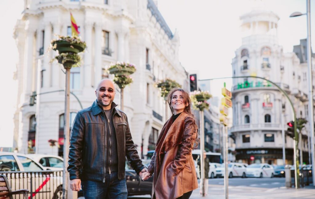 Pareja sonriente de la mano en el inicio de la Gran Vía de Madrid, con el Edificio Grassy y su icónico cartel de Rolex al fondo
