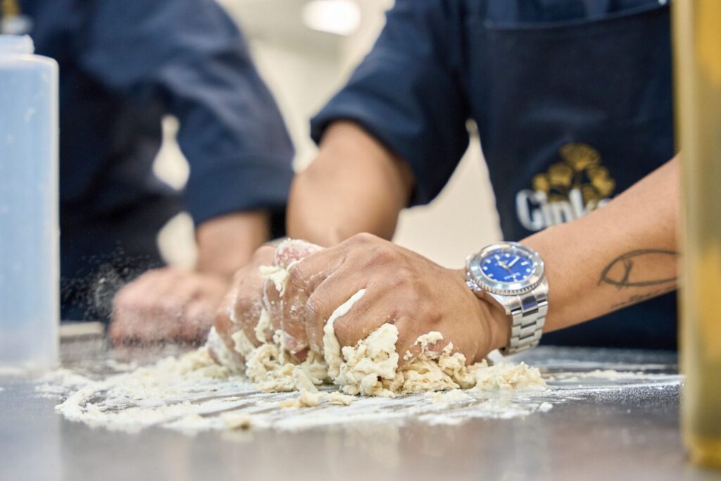 Primer plano de un cocinero con delantal de Ginkgo amasando manualmente sobre una mesa de acero inoxidable, con harina esparcida y un reloj de esfera azul destacado