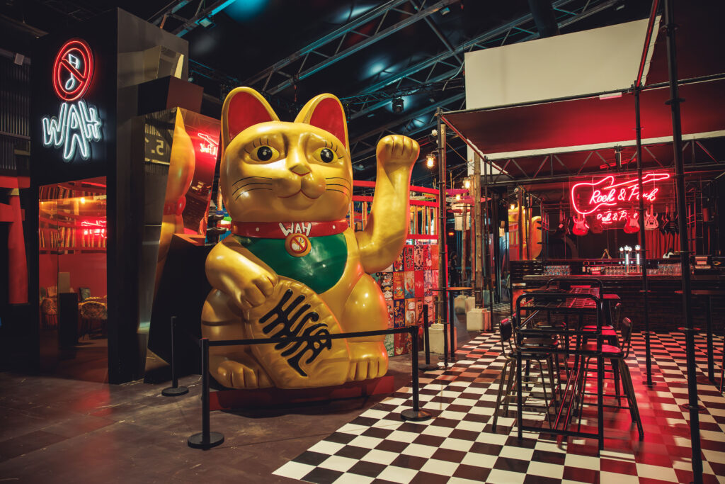 Estatua gigante de un Maneki-neko (gato de la fortuna) dorado con el logo de WAH en el collar, situado en el Food Hall junto a un neón rojo de 'Rock & Roll' y suelo de cuadros blancos y negros