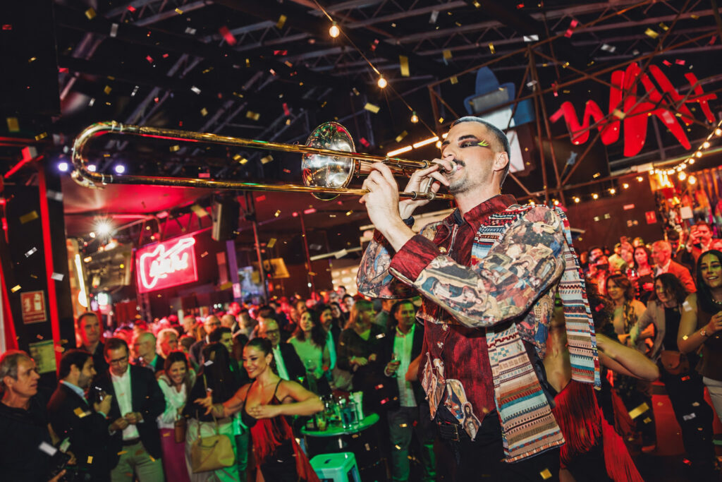 Músico con maquillaje artístico tocando el trombón en el Food Hall de WAH Show, rodeado de público y confeti, con el logotipo de WAH en neón rojo al fondo.