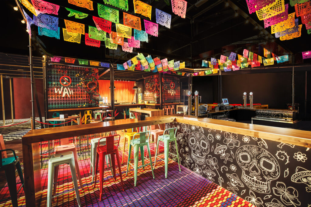 Área temática mexicana de WAH Show decorada con guirnaldas de papel picado, ilustraciones de calaveras mexicanas en la barra y logotipos de WAH con la nota musical tachada