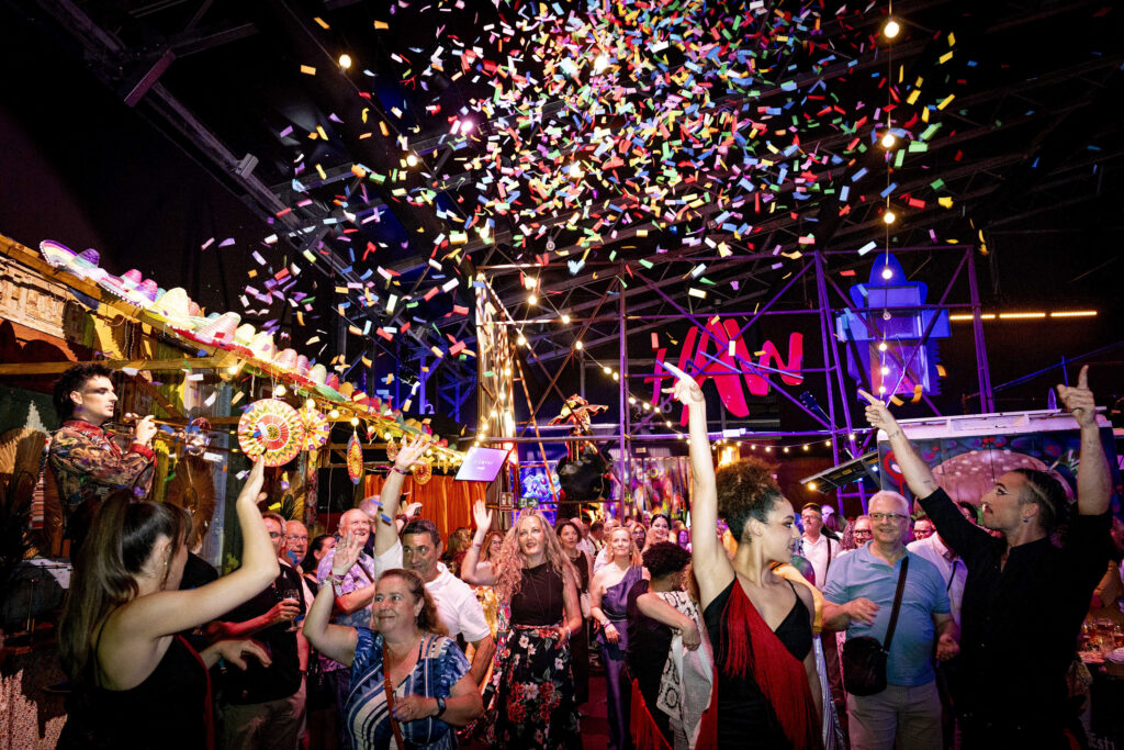 Público diverso celebrando y bailando bajo una gran lluvia de confeti de colores en el Food Hall de WAH Show, con el logotipo rojo de WAH iluminado al fondo