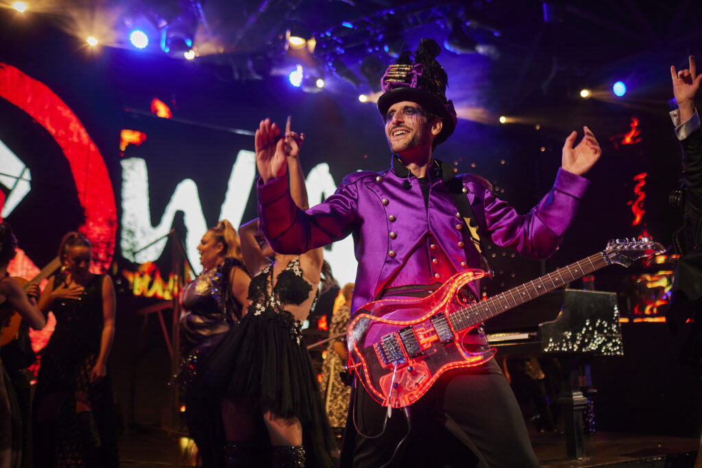 Músico de WAH Show con chaqueta militar púrpura y chistera steampunk tocando una guitarra eléctrica transparente con luces LED rojas, con bailarines y fuego en las pantallas al fondo.