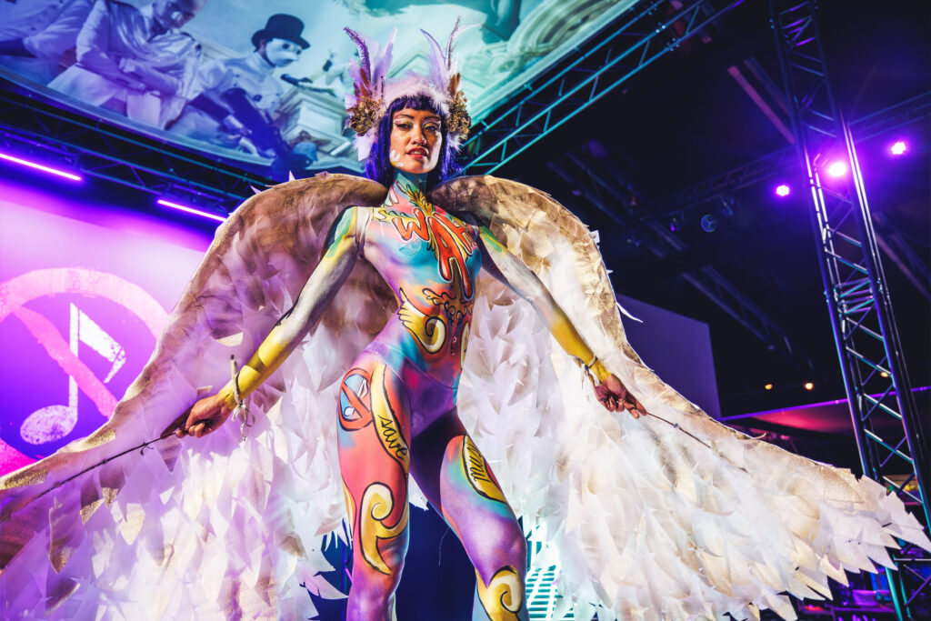 Artista de WAH Show con el cuerpo cubierto por body paint multicolor estilo grafiti con la palabra 'WAH' en el pecho, luciendo grandes alas de plumas blancas y doradas