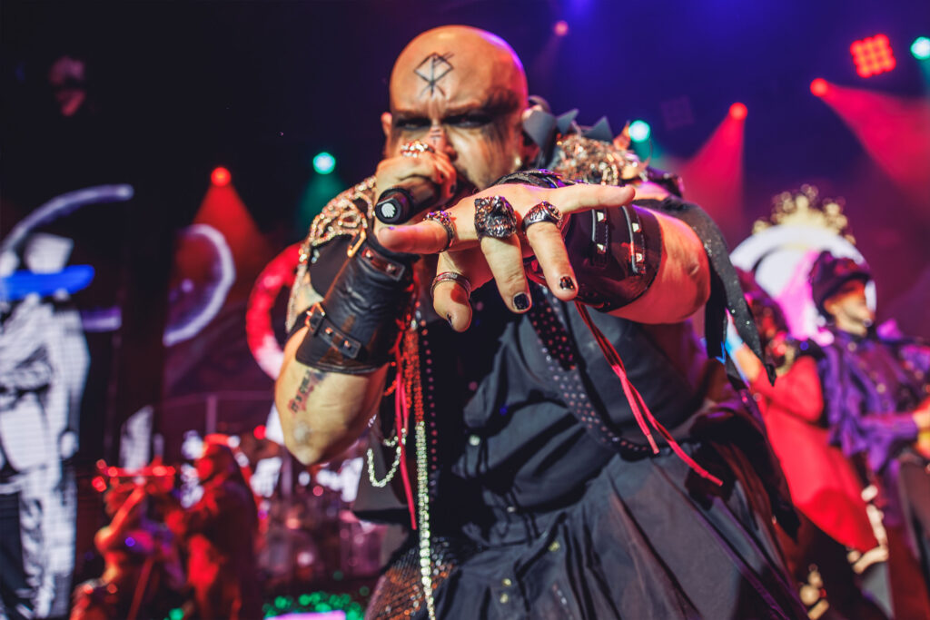 Cantante solista de WAH Show con una runa tatuada en la frente, maquillaje oscuro en los ojos y múltiples anillos de calavera, señalando a la cámara mientras canta bajo luces de escenario rojas y verdes