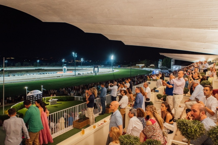 Público en las terrazas del Hipódromo de la Zarzuela en Madrid durante un evento nocturno, bajo los icónicos voladizos de hormigón y con publicidad de Santander junto a la pista iluminada.