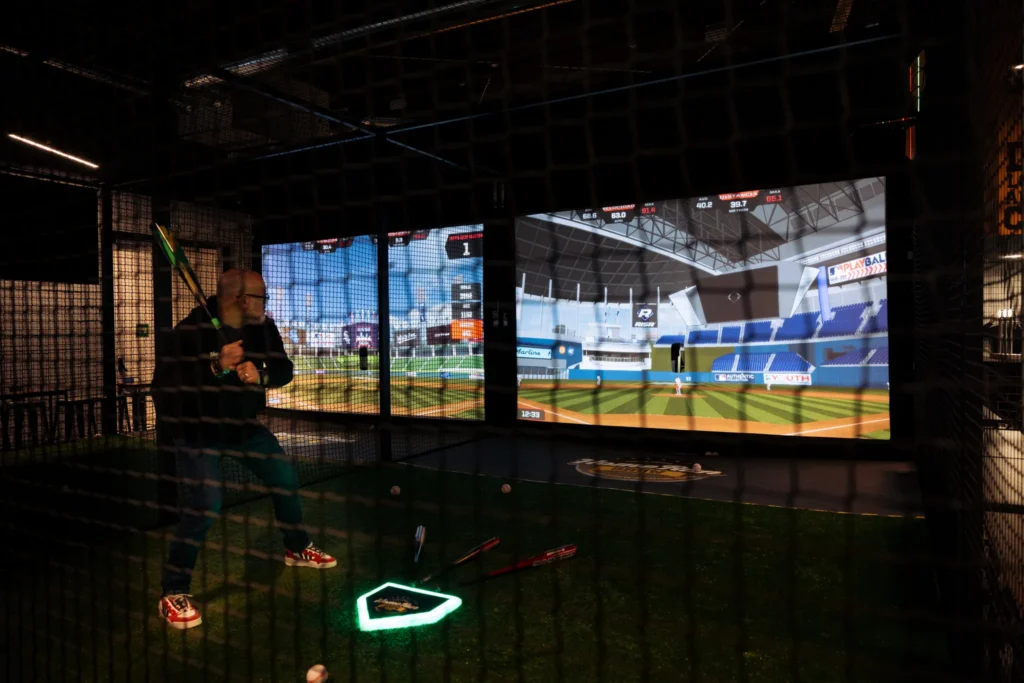 Hombre bateando en un simulador de béisbol interactivo de Home Run Social con pantallas gigantes que proyectan un estadio virtual y estadísticas de velocidad y distancia en tiempo real.