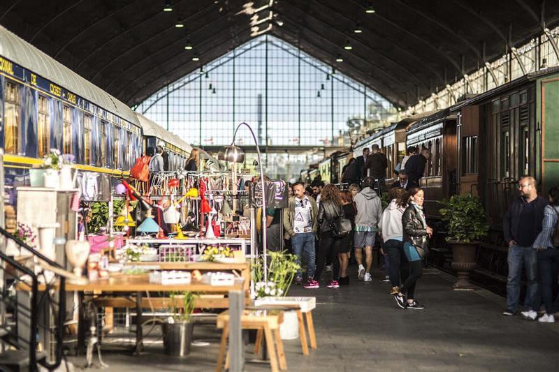Visitantes en el Mercado de Motores dentro del Museo del Ferrocarril de Madrid, con puestos de muebles y lámparas vintage situados entre vagones de tren históricos bajo la gran bóveda de hierro y cristal.