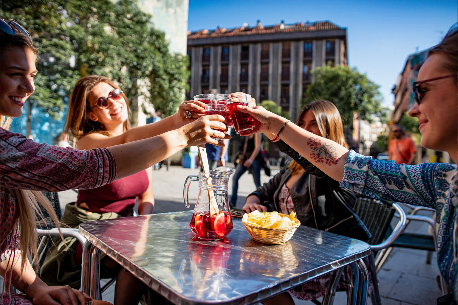 Cuatro jóvenes brindando con copas de sangría en una terraza soleada de Madrid, con una jarra con frutas y un cuenco de patatas fritas sobre una mesa metálica
